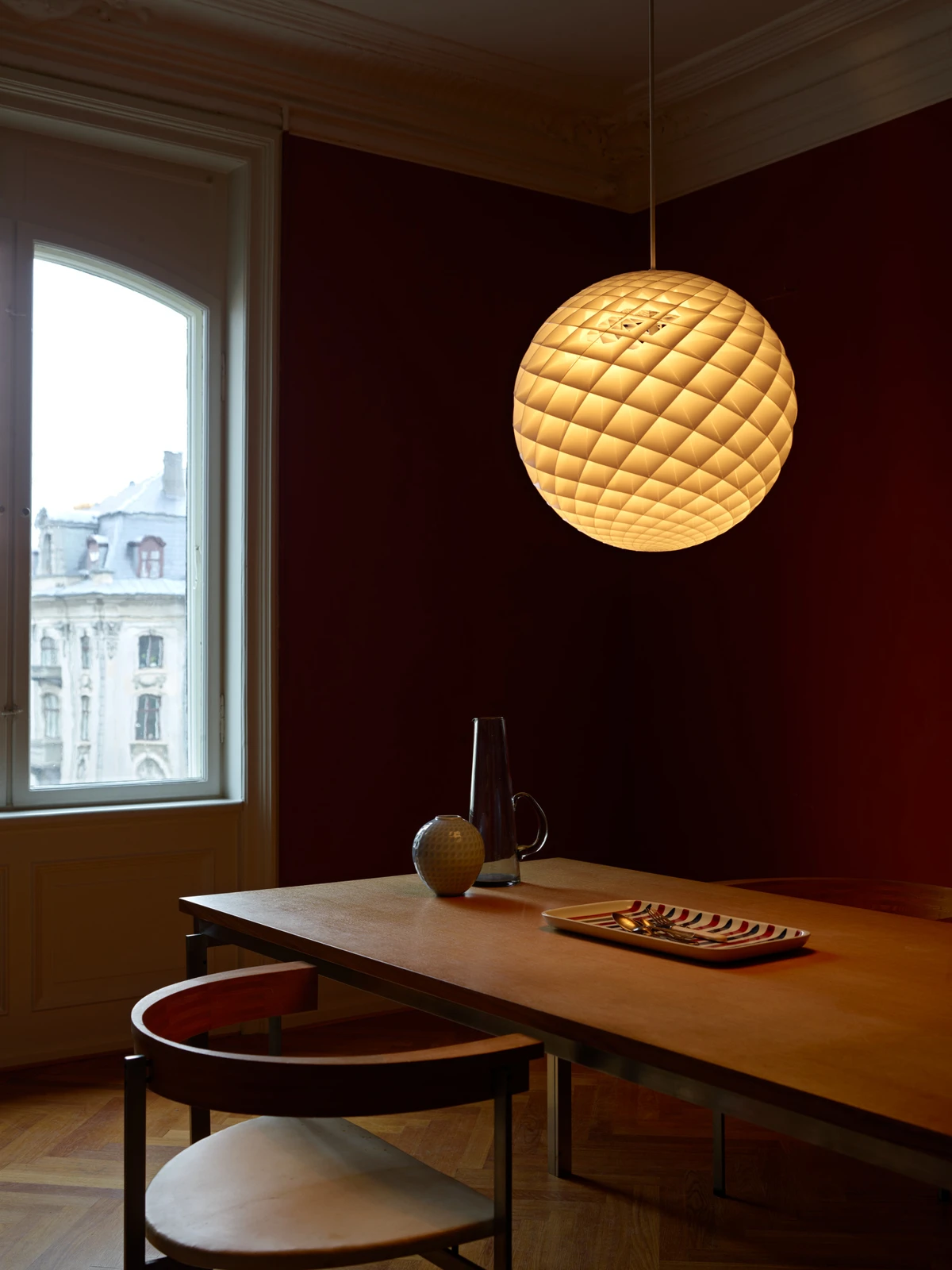 Een warm gloeiende Patera hanglamp hangt boven een eettafel in een kamer met donkerrode muren. © 2026 Louis Poulsen.