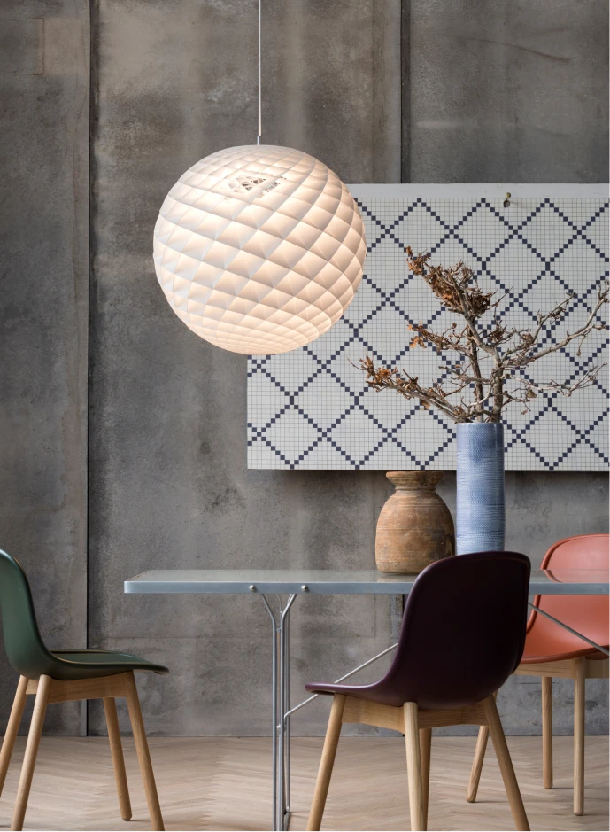 Een witte Patera hanglamp verlicht een eettafel met kleurrijke stoelen tegen een onafgewerkte betonnen muur. © 2026 Louis Poulsen.