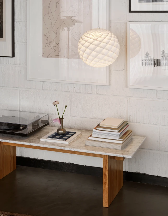 Een brandende Patera-hanglamp hangt boven een marmeren tafel in een lichte ruimte met witte muren en ingelijste kunst. © 2026 Louis Poulsen.