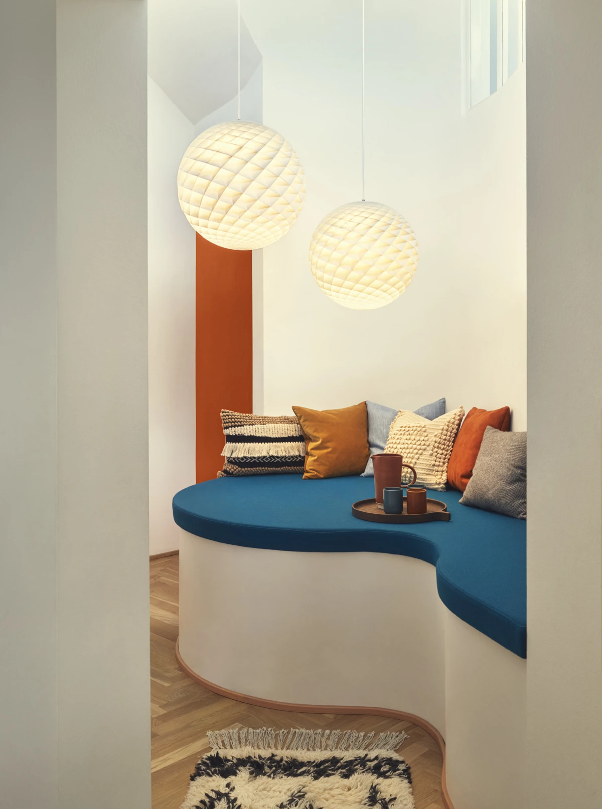 Two white Patera Pendant lights glow above a built-in bench with blue upholstery and colourful cushions. © 2026 Louis Poulsen.