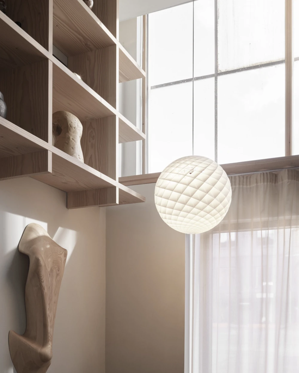 Een witte Patera Pendant hangt naast een groot raam in een lichte ruimte met houten planken in een lichte tint. © 2026 Louis Poulsen.
