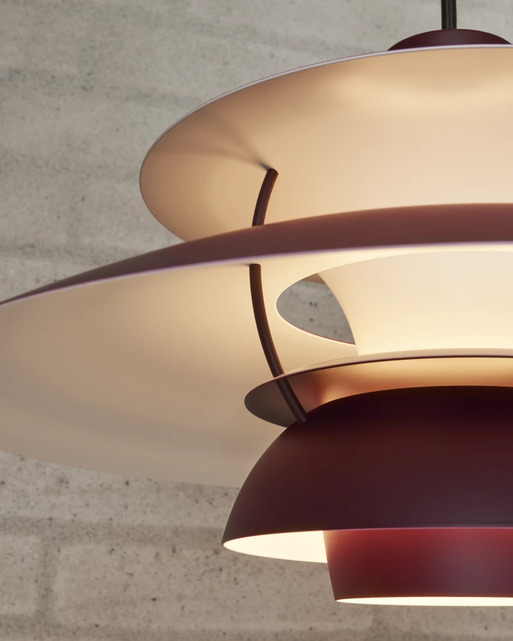 Close-up van een bordeauxrode PH 5 hanglamp met warm verlichte kappen tegen een ruwe betonnen muur. © 2026 Louis Poulsen.