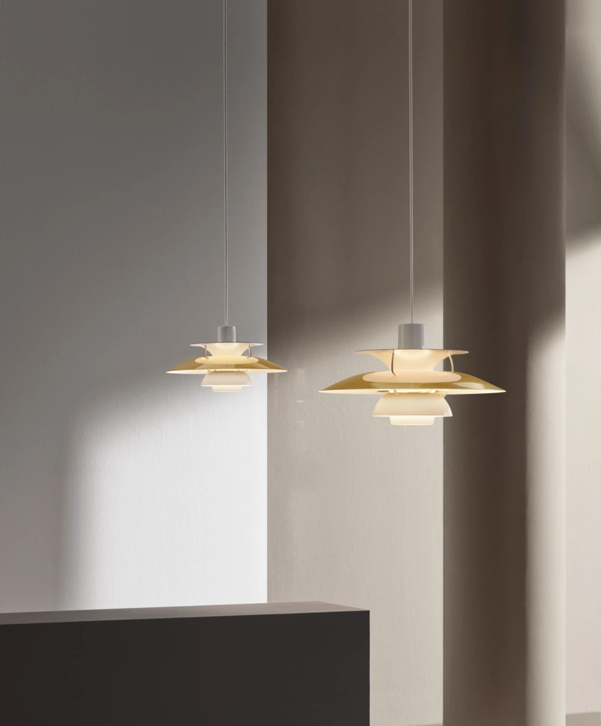 Twee PH 5-hanglampen in verschillende formaten hangen voor een lichte muur en geven warm licht. © 2026 Louis Poulsen.