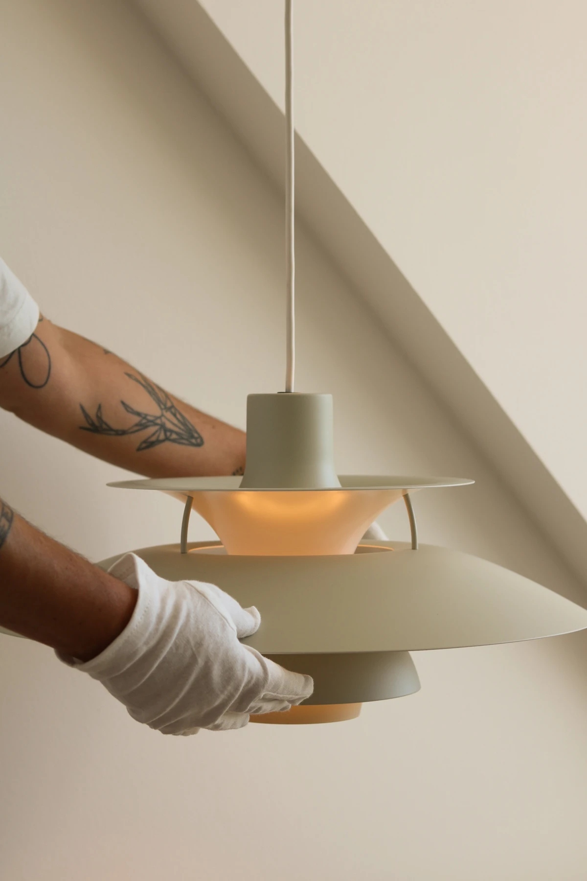 Een lichtgroene PH 5-pendel brandt terwijl iemand hem bijstelt voor een beige muur. © 2026 Louis Poulsen.