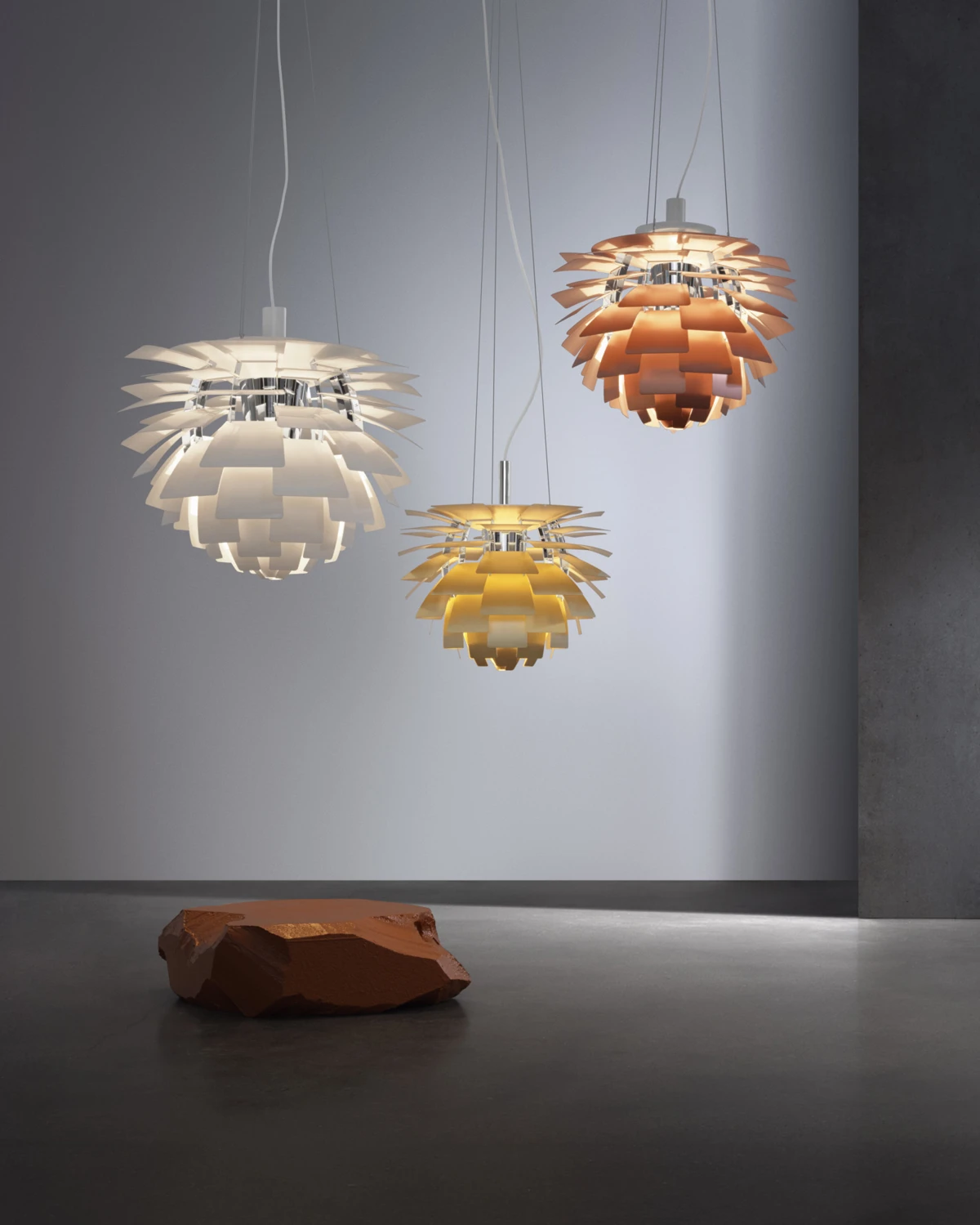 Drie PH Artichoke-pendels in wit, messingkleurig en koper hangen tegen een grijze muur. © 2026 Louis Poulsen.