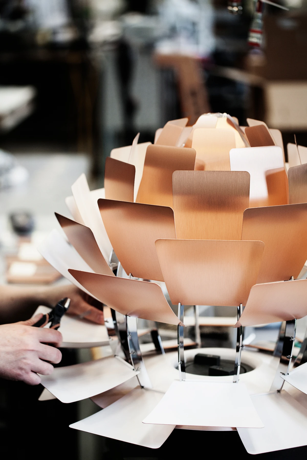 Een PH Artichoke hanglamp met koperkleurige lamellen wordt in een werkplaats gemonteerd. © 2026 Louis Poulsen.