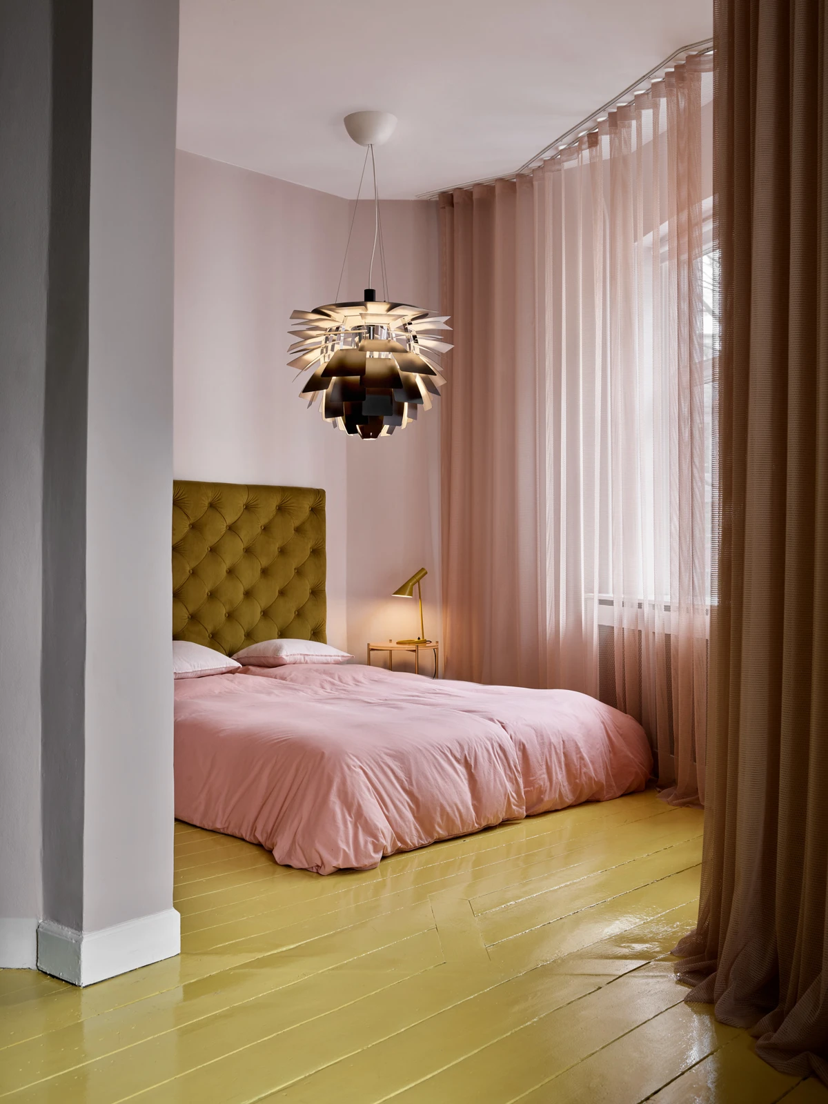Een zwarte PH Artichoke hanglamp en een gele AJ Mini tafellamp in een slaapkamer met roze beddengoed en een okergele vloer. © 2026 Louis Poulsen.