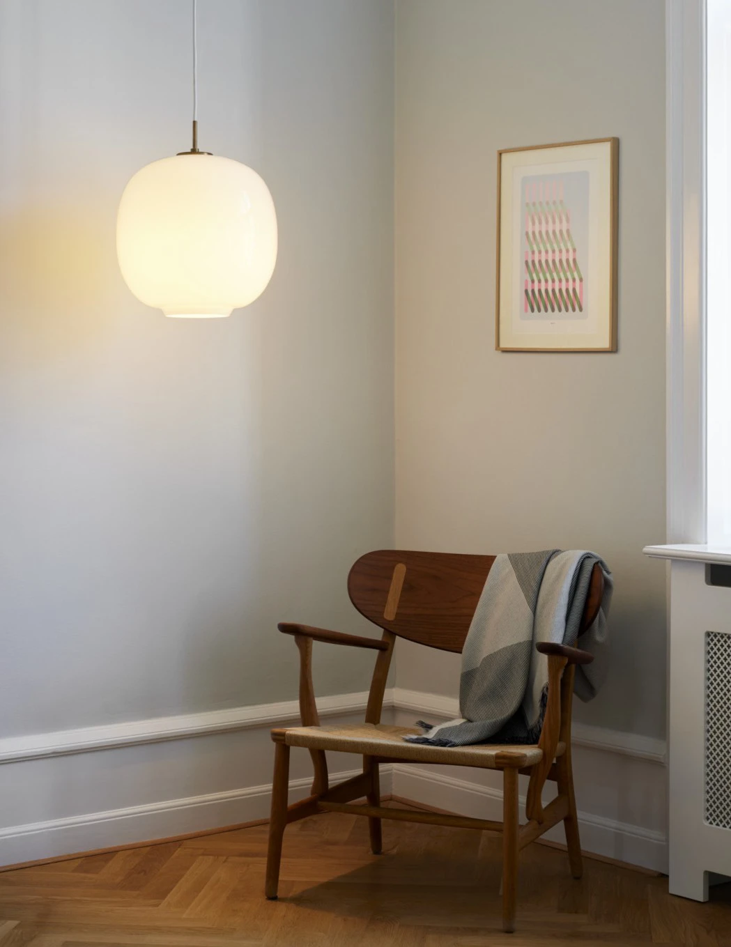 Een opaalwitte VL 45 Radiohus hanglamp verlicht een houten stoel in een rustige kamer met visgraatparket. © 2026 Louis Poulsen.