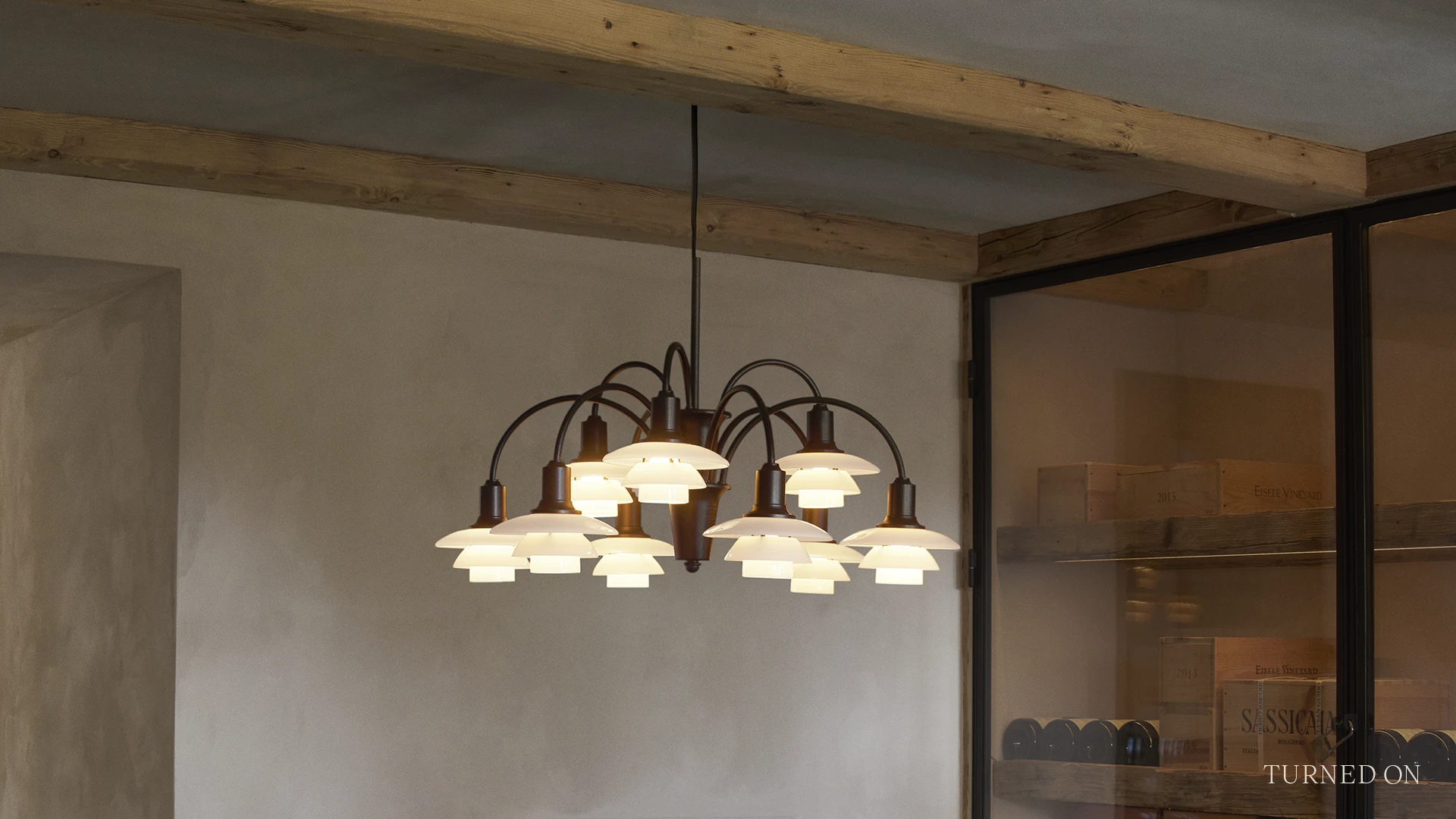 De PH 1/1 Chandelier Centenary Edition verlicht een ruimte met zichtbare houten balken en een glazen wand. © 2026 Louis Poulsen.