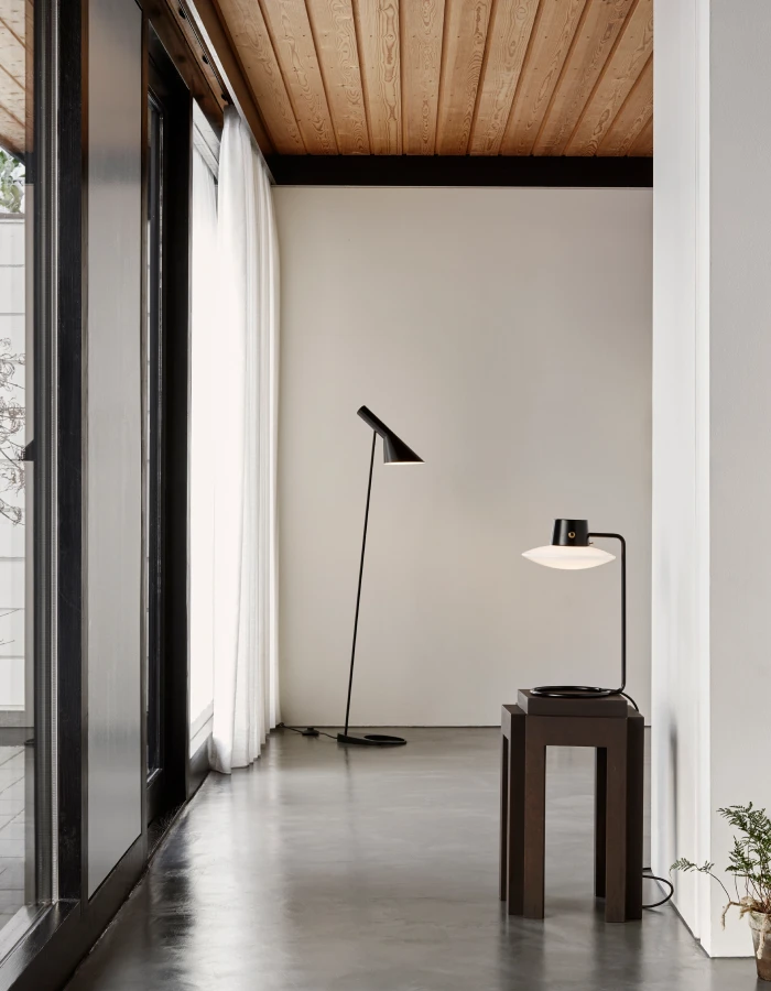 En sort AJ Floor Lamp og en AJ Oxford bordlampe i et lyst rum med trægulv og loft. © 2026 Louis Poulsen.