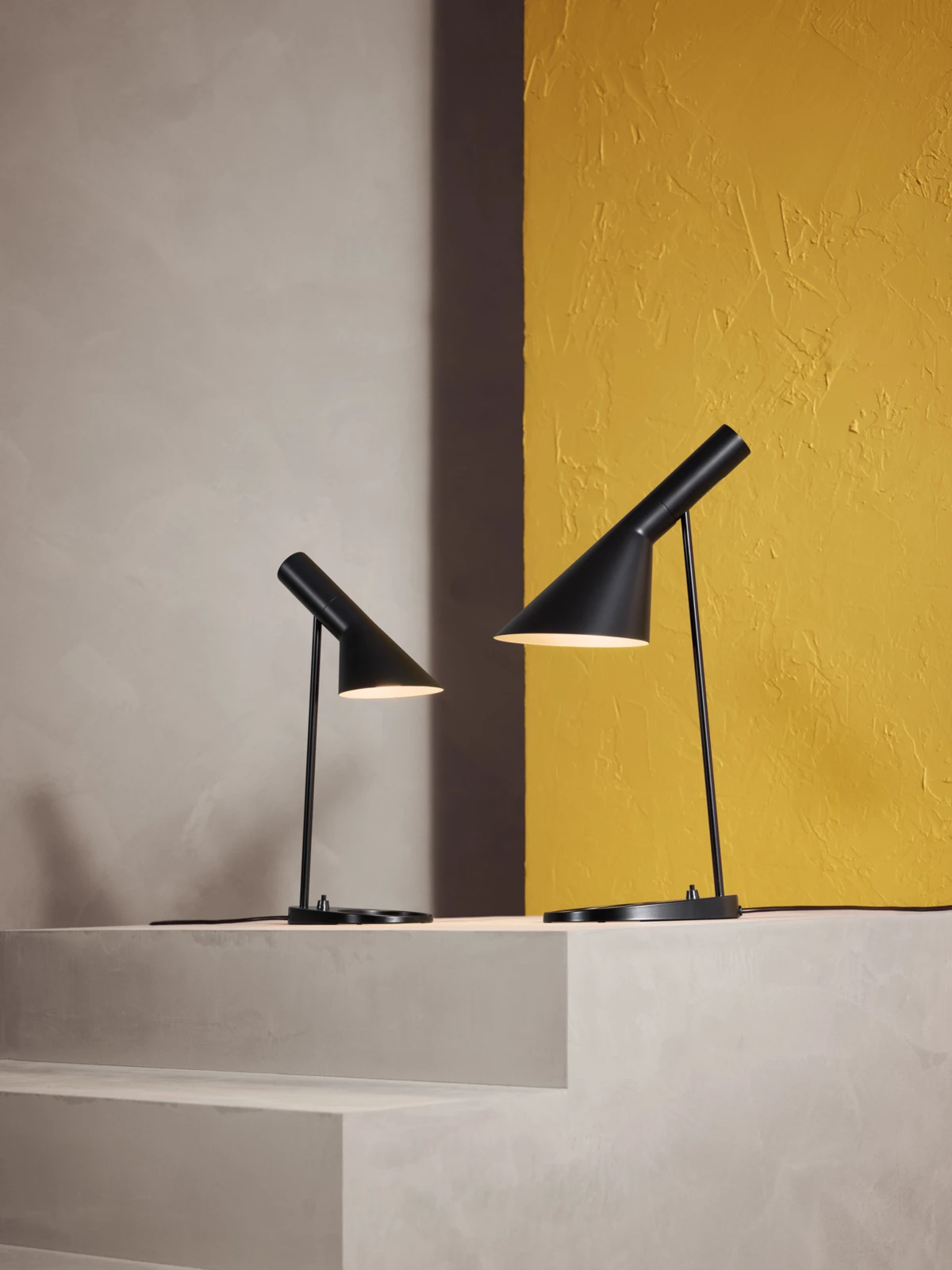 AJ Mini Table lamp & AJ Table lamp in black on concrete steps with a yellow wall backdrop, © 2025 Louis Poulsen.