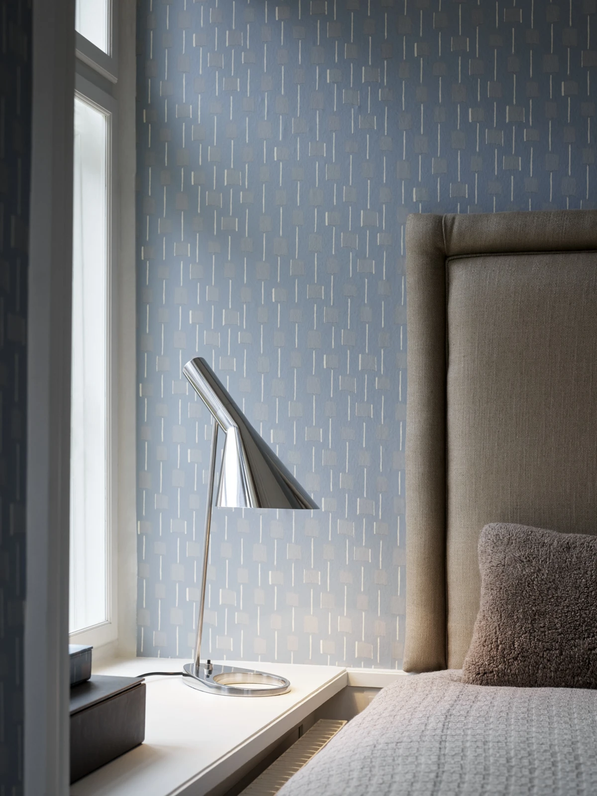A chrome AJ Table Lamp stands on a bedside table beside a bed against blue patterned wallpaper. © 2026 Louis Poulsen.