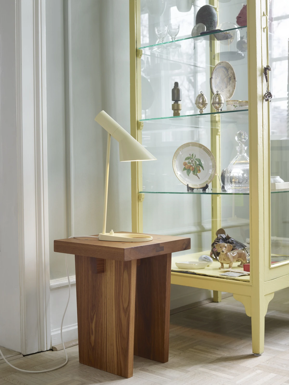 A soft lemon AJ Mini Table Lamp stands on a small wooden table beside a glass cabinet in a bright Frederiksberg interior. © 2026 Louis Poulsen.