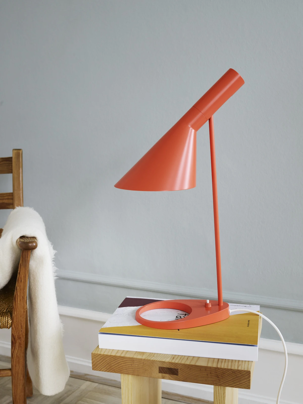 An orange AJ Table Lamp rests on books atop a small wooden table against a pale grey wall. © 2026 Louis Poulsen.