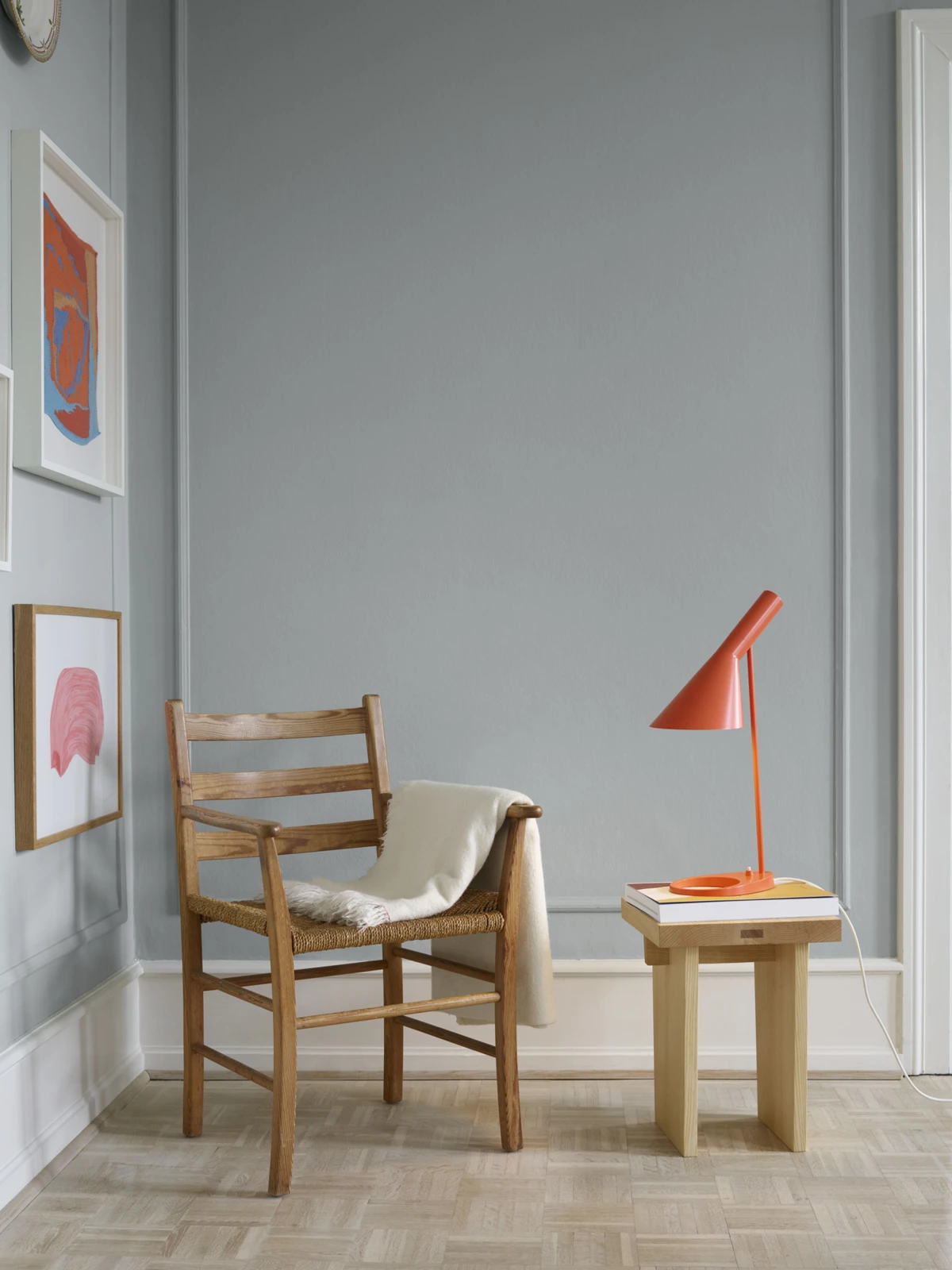 An electric orange AJ Table Lamp sits on a small wooden side table beside a wooden chair in a light-filled room. © 2026 Louis Poulsen.