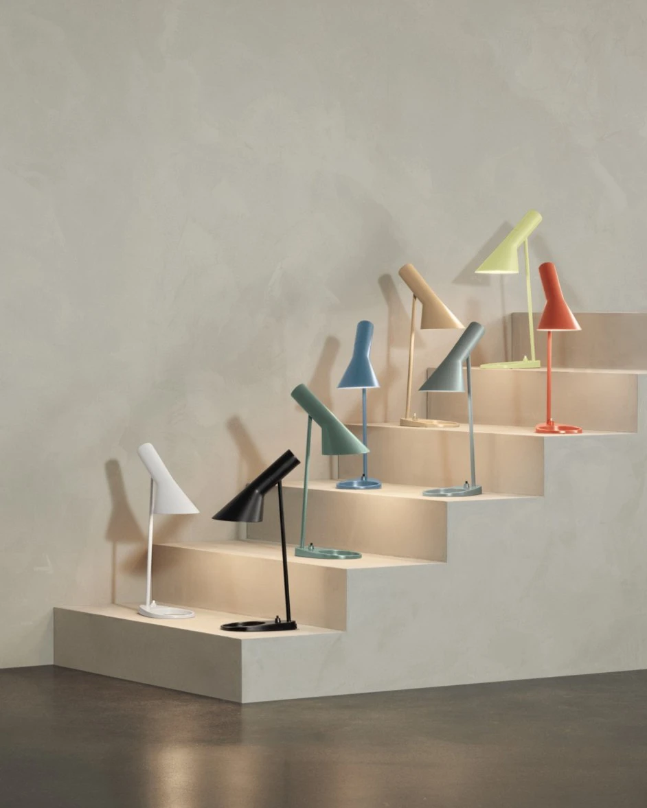 AJ Table Lamps in several colours stand on pale stepped plinths against a textured wall. © 2026 Louis Poulsen.