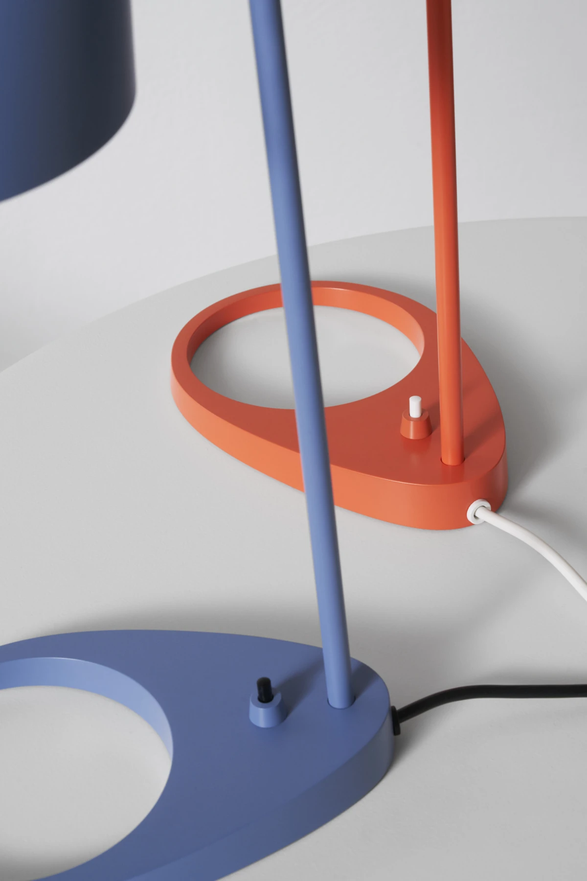 Two AJ Table Lamps in blue and orange sit on a white surface, shown close up with their distinctive circular bases. © 2026 Louis Poulsen.