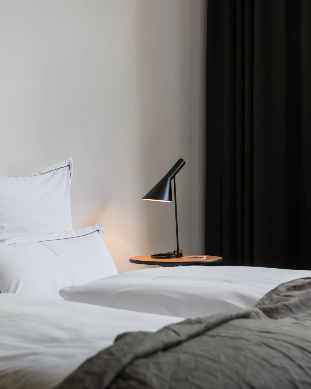 A black AJ Table Lamp on a round bedside table beside a bed with white linens. © 2026 Louis Poulsen.