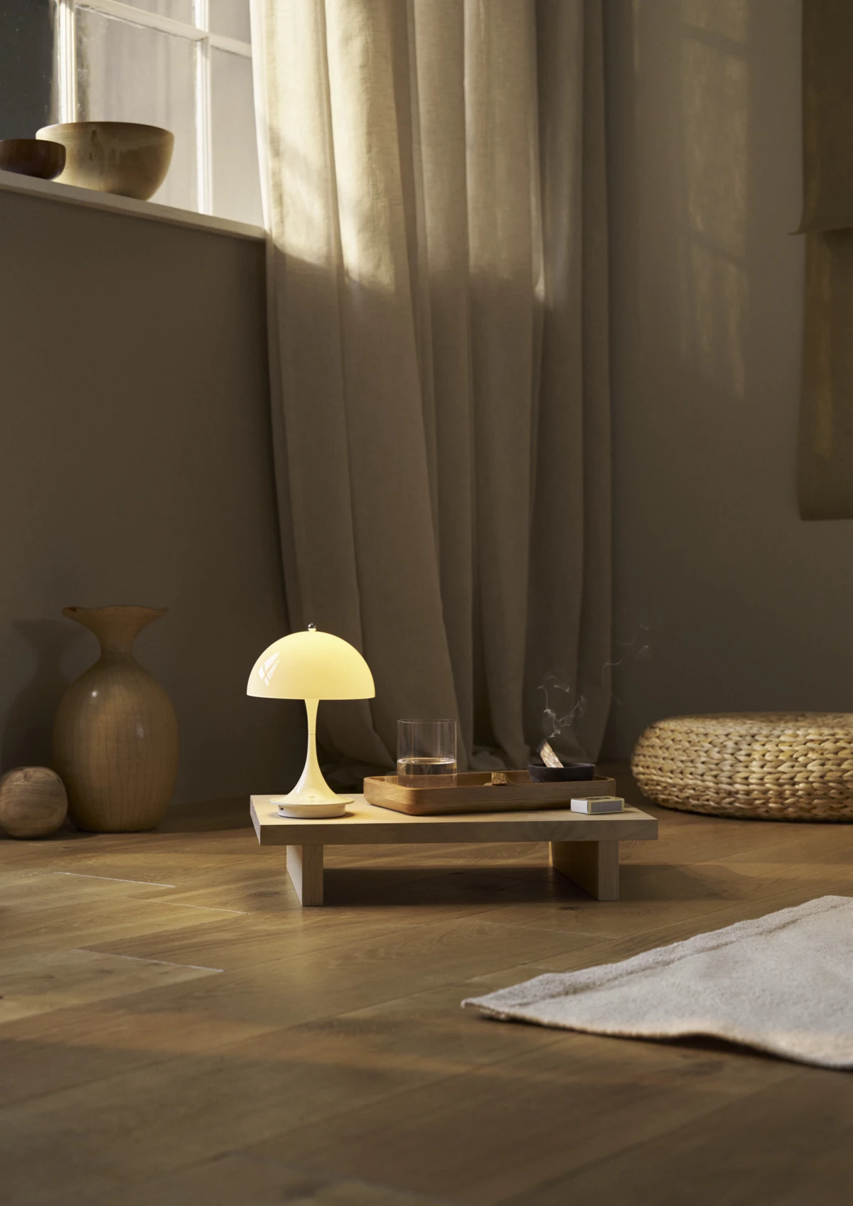 Een beige Panthella 160 Portable Lamp schijnt op een lage houten tafel in een woonkamer met lichte gordijnen en een houten vloer. © 2026 Louis Poulsen.