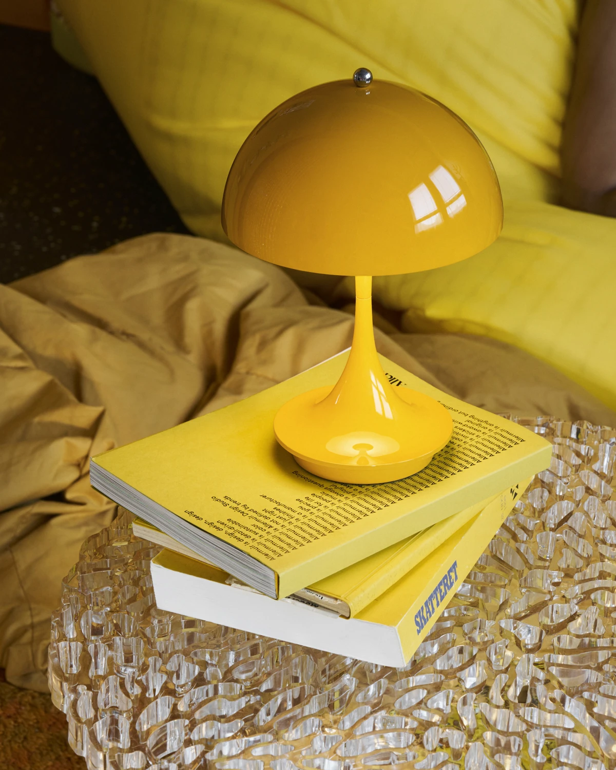 Een gele Panthella 160 Portable Lamp staat op boeken op een transparante tafel naast een bank. © 2026 Louis Poulsen.