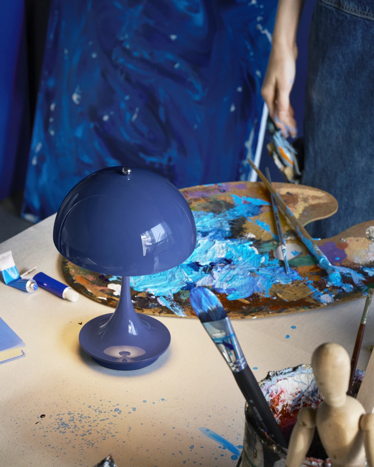 Een donkerblauwe Panthella 160 Portable tafellamp staat tussen penselen en een palet vol verf op een werktafel van een kunstenaar. © 2026 Louis Poulsen.