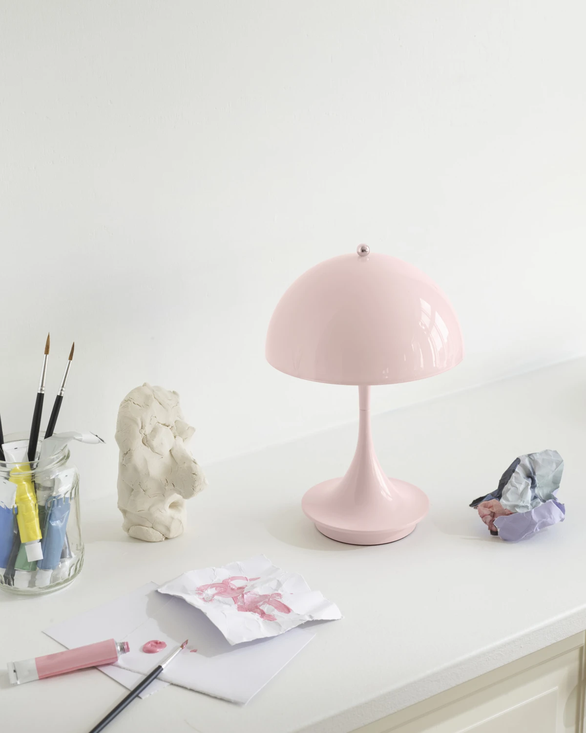 Een Panthella 160 draagbare lamp in Pale Rose staat op een wit bureau tussen verf, penselen en tekeningen. © 2026 Louis Poulsen.