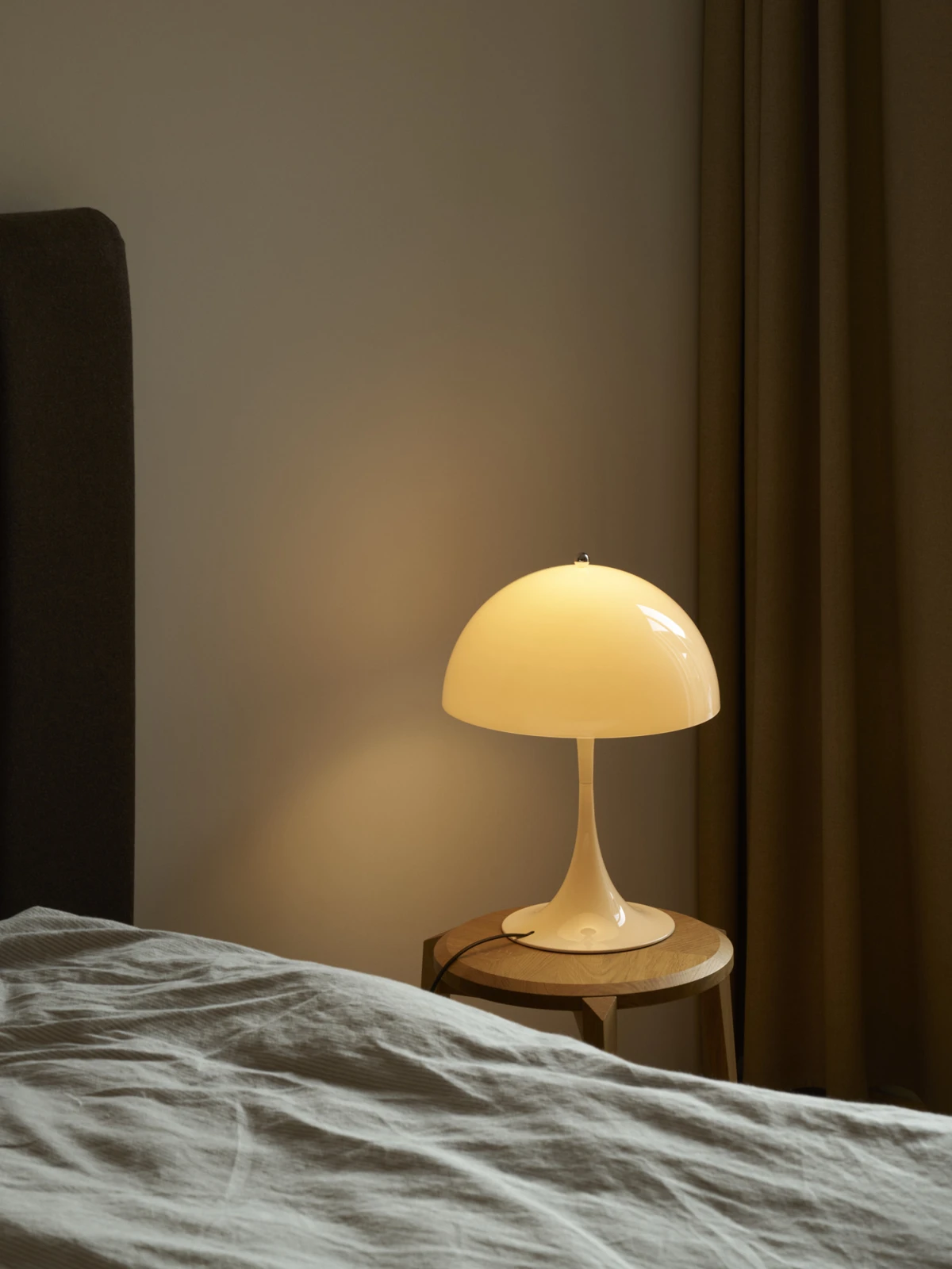 A beige Panthella 320 Table Lamp casts a warm glow from a wooden bedside table. © 2026 Louis Poulsen.