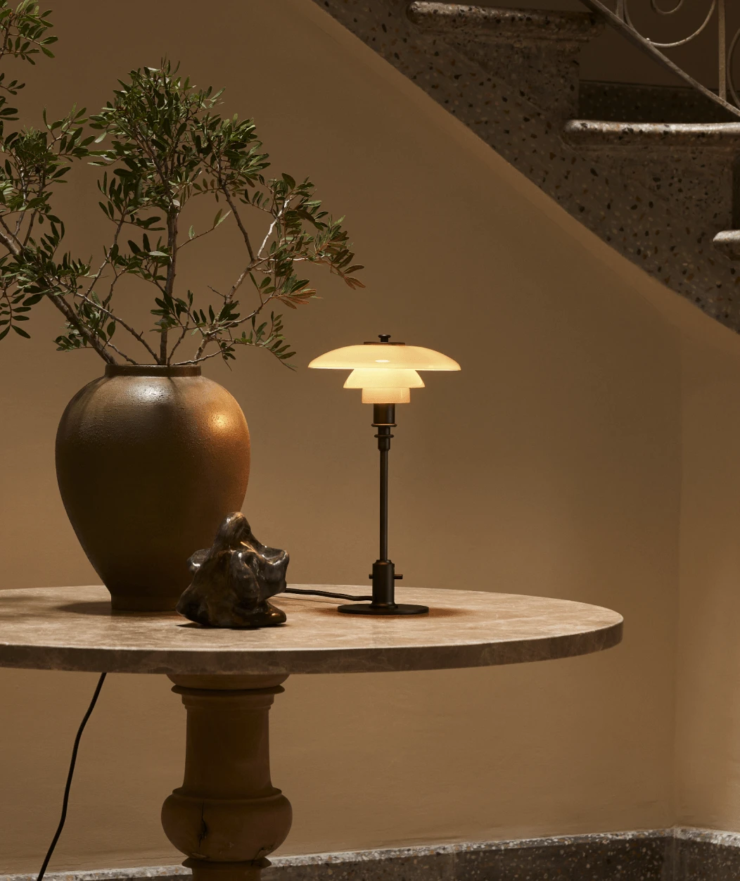 De PH 1/1 Portable Lamp met opaalwitte, verlichte kappen staat op een ronde stenen tafel naast een trap. © 2026 Louis Poulsen.