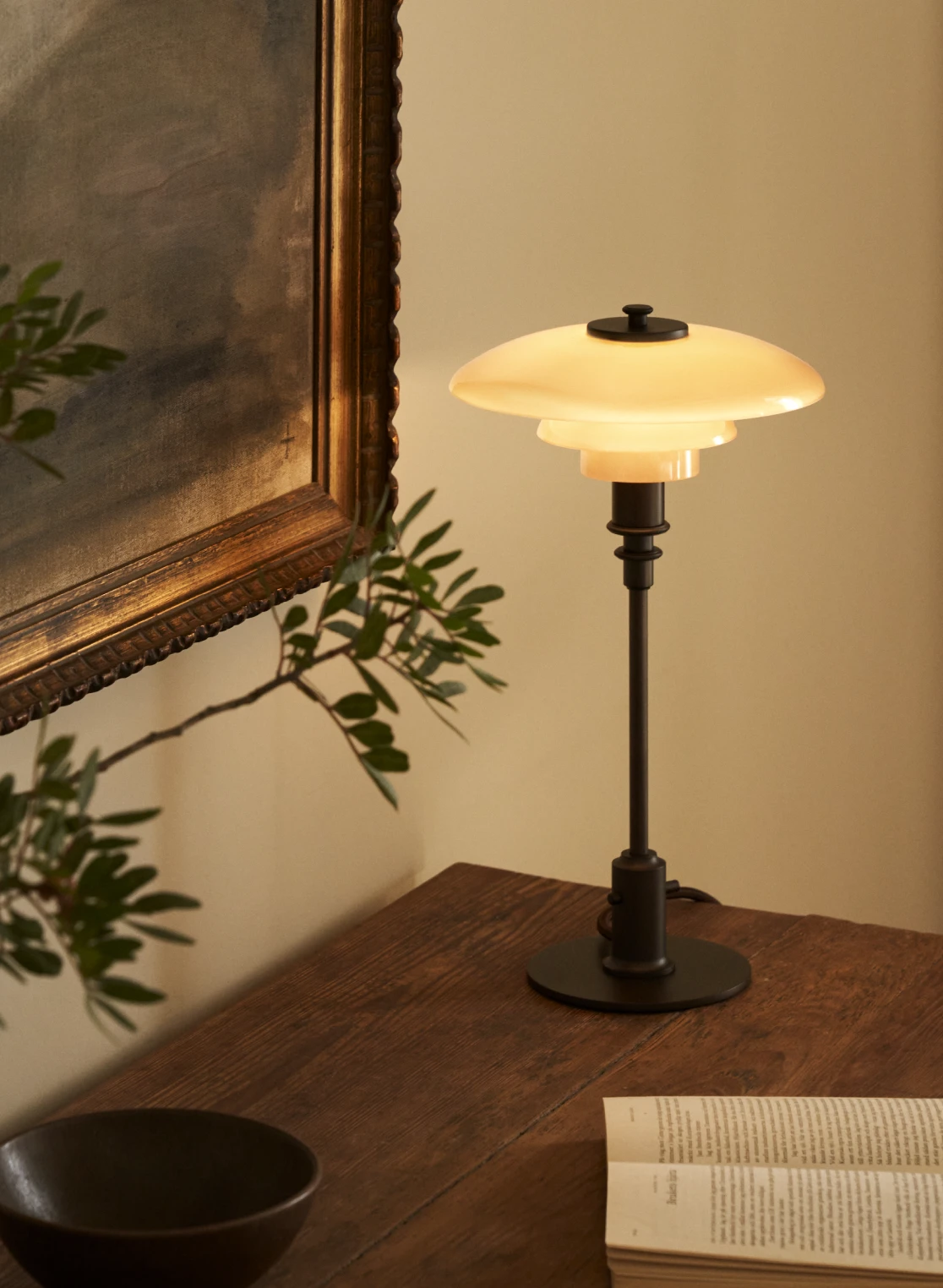 A PH 2/1 Dusty Terracotta Table Lamp casts a warm glow on a wooden table beside a book and framed painting. © 2026 Louis Poulsen.
