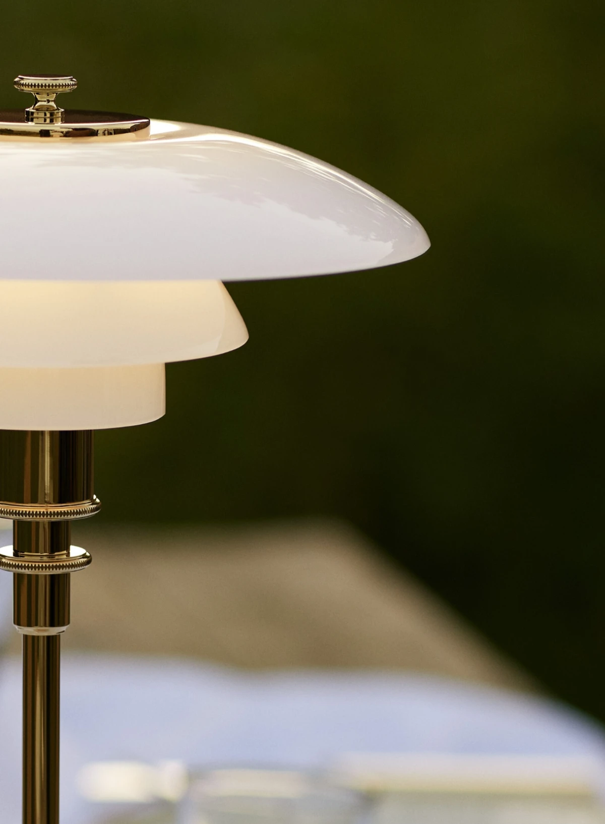 De draagbare PH 2/1 lamp met opaalwitte kappen en messingkleurige staander geeft warm licht tegen een donkergroene achtergrond. © 2026 Louis Poulsen.