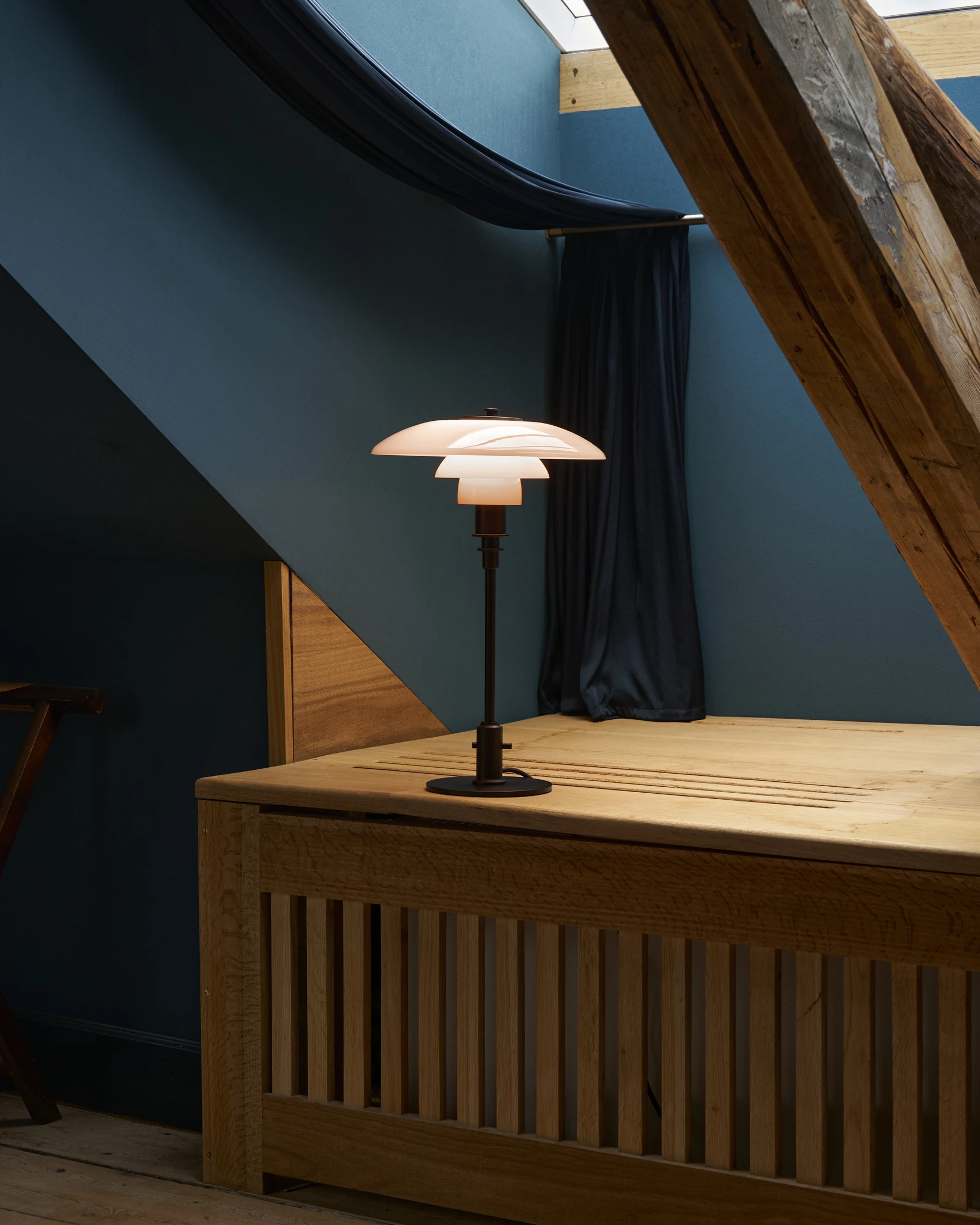 A terracotta-shaded PH 3/2 Table Lamp glows on a wooden table in a room with dark blue walls. © 2026 Louis Poulsen.