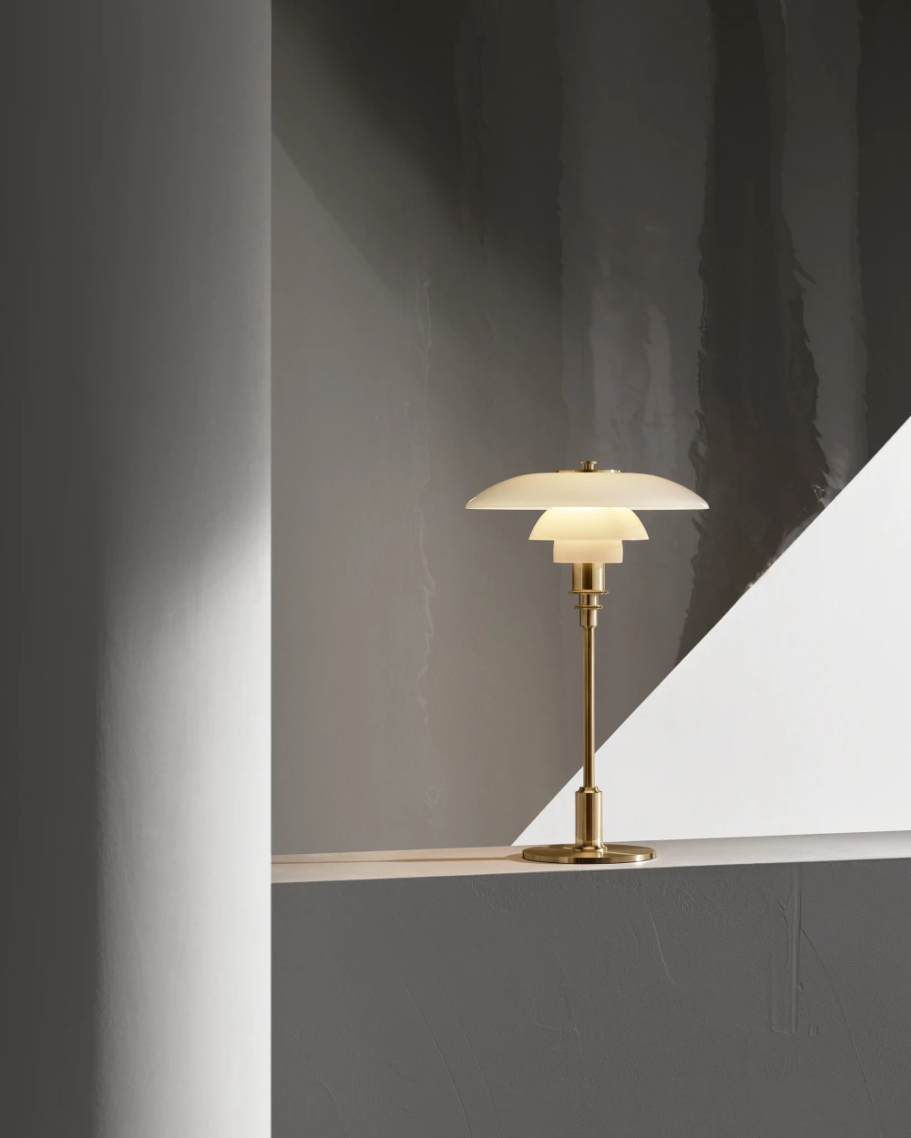 PH 3/2 Brass Opal Glass bordlampe i messing og opalglas står på en afsats mod en mørk væg. © 2026 Louis Poulsen.