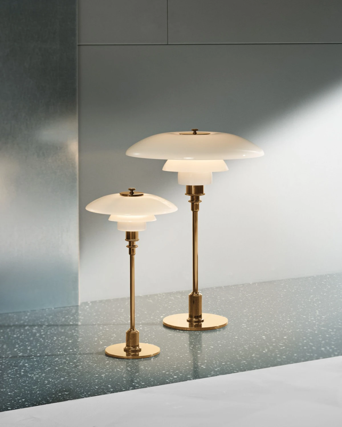 Una lampada da tavolo PH 2/1 Aged Brass e una PH 3½-2½ in ottone, con paralumi bianco opalino, sono appoggiate su un piano. © 2026 Louis Poulsen.