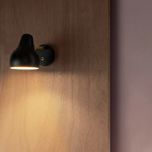 Black VL 38 Wall Lamp with a brass detail on a wooden wall, casting a warm pool of light. © 2026 Louis Poulsen.