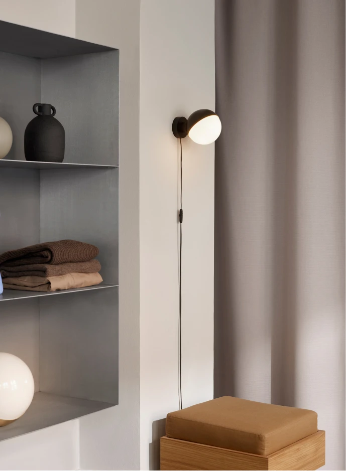 Eine schwarze VL Studio Wall Wandleuchte mit Opalglasschirm leuchtet neben einem Einbauregal und einer Holzbank. © 2026 Louis Poulsen.