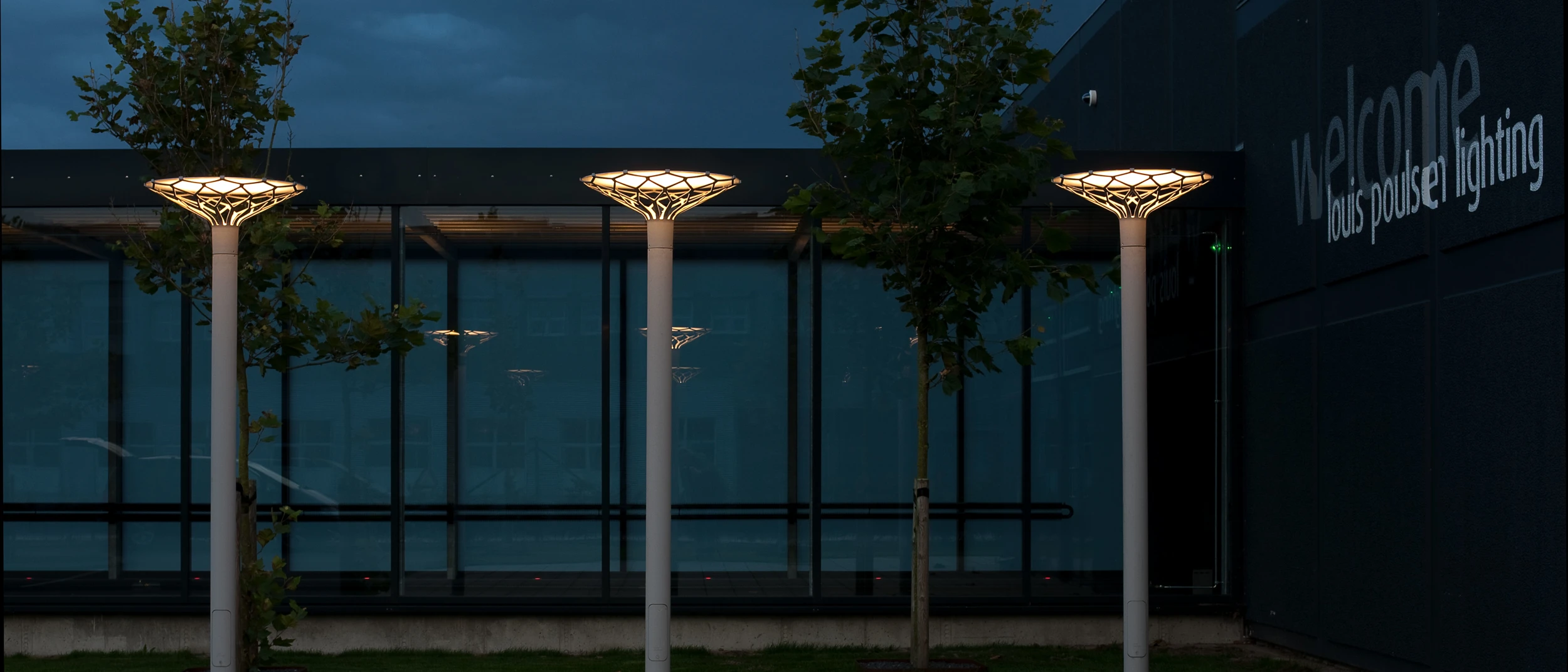 Three Flindt Plaza bollards glow outside Louis Poulsen’s glass-fronted building at dusk. © 2026 Louis Poulsen.