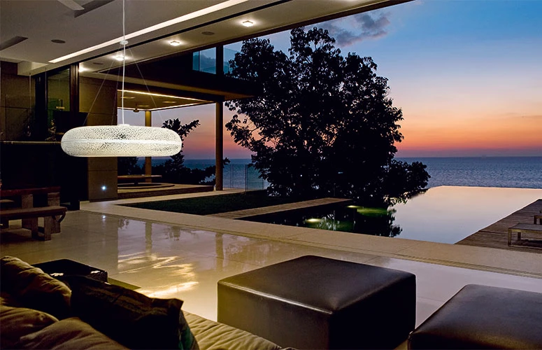 An elongated pendant light hangs in a modern living room overlooking the sea at sunset. © 2026 Louis Poulsen.