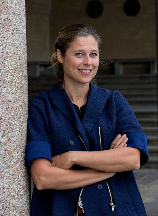 A woman in a dark blue jacket stands with her arms crossed beside a stone wall. © 2026 Louis Poulsen.