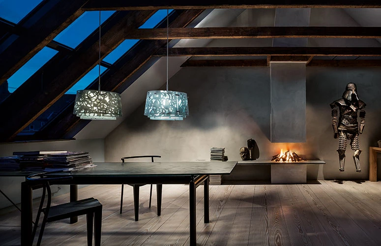 Two Collage Pendants hang above a dining table in a dark attic room with exposed timber beams and a fireplace. © 2026 Louis Poulsen.