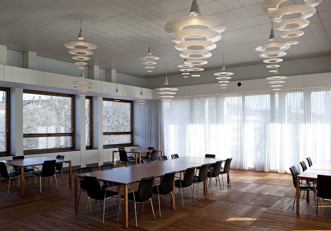 White Enigma pendants hang above wooden tables in a bright meeting and dining room with large windows. © 2026 Louis Poulsen.