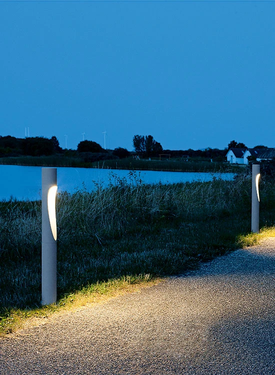 Flindt Bollard bollards illuminate a gravel path beside the water at dusk, surrounded by tall grasses. © 2026 Louis Poulsen.
