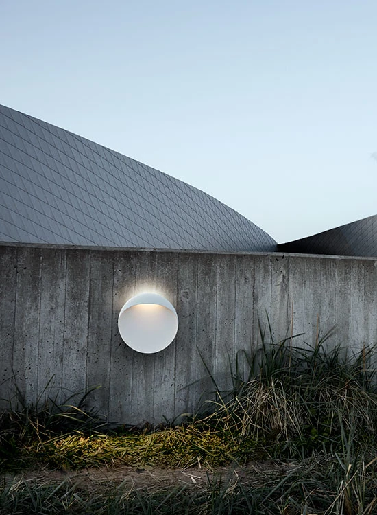 A white Flindt Wall Lamp glows on a concrete wall beside a modern building and ornamental grasses. © 2026 Louis Poulsen.