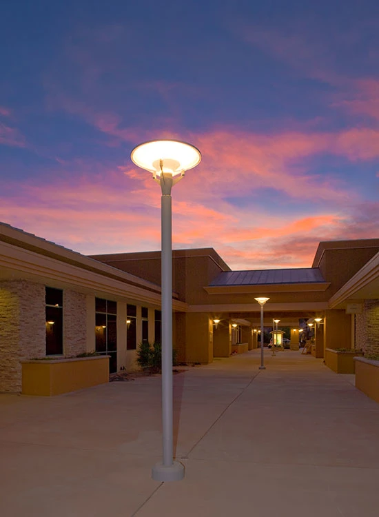 A Kipp Post illuminates a walkway between low buildings beneath a colourful sunset sky. © 2026 Louis Poulsen.