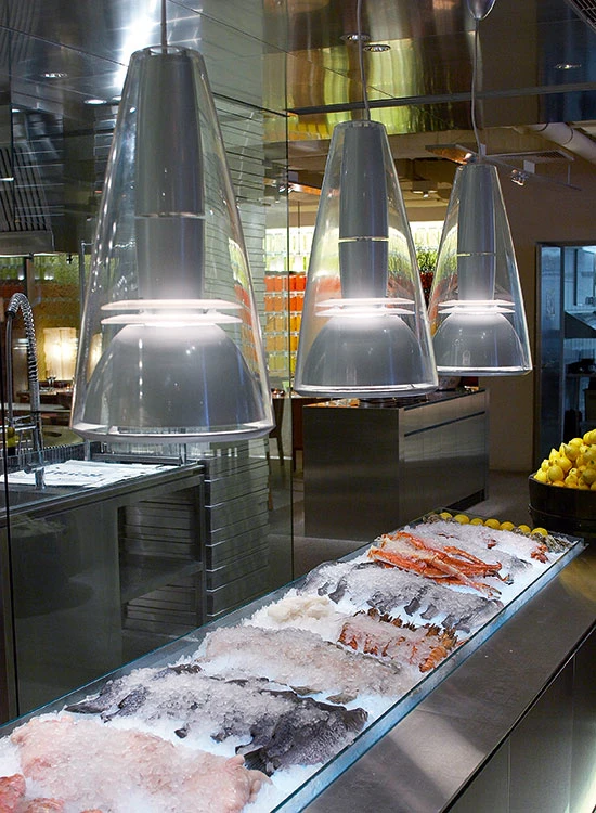 Three translucent pendant lights illuminate a fish counter in a modern room with stainless-steel surfaces. © 2026 Louis Poulsen.