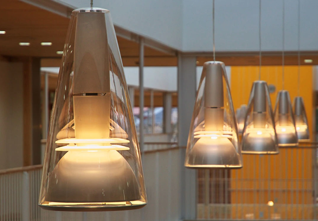 Louis Poulsen pendants with clear, tapered outer shades cast a warm glow in a row in a modern atrium. © 2026 Louis Poulsen.