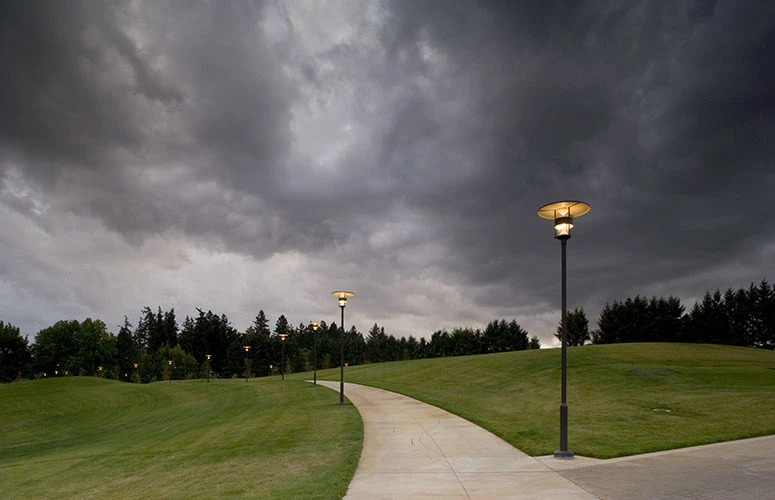 Louis Poulsen outdoor post lights line a curving path through a green park beneath dark clouds. © 2026 Louis Poulsen.