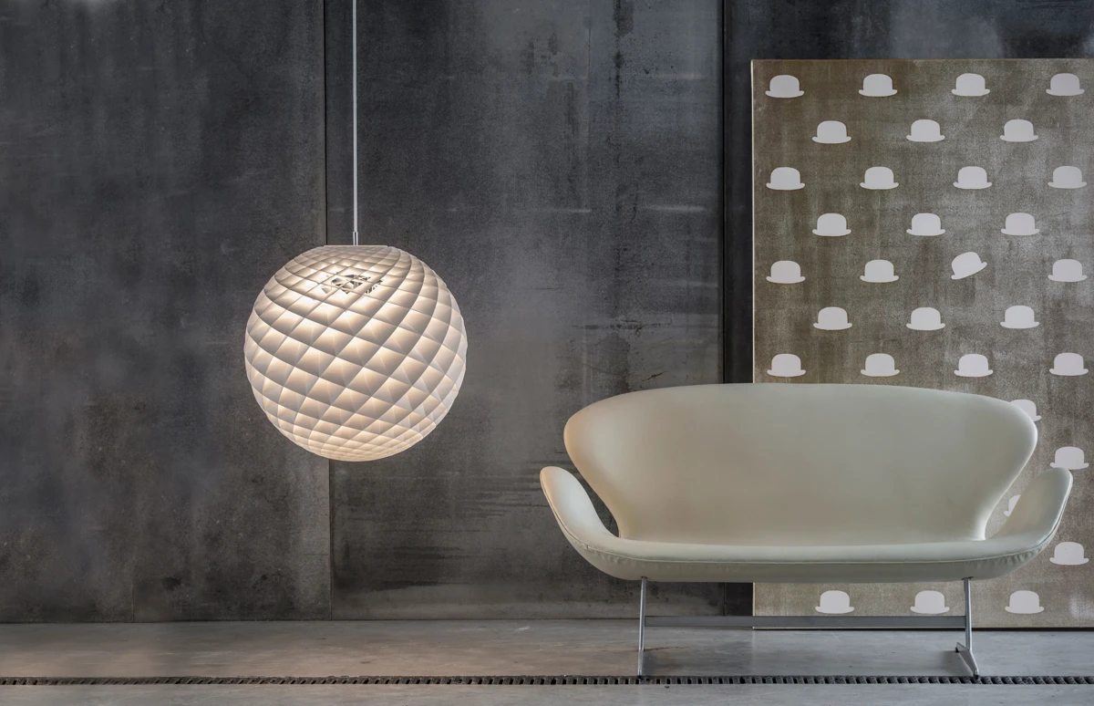 An illuminated Patera Pendant hangs beside a pale sofa against a dark concrete wall. © 2026 Louis Poulsen.