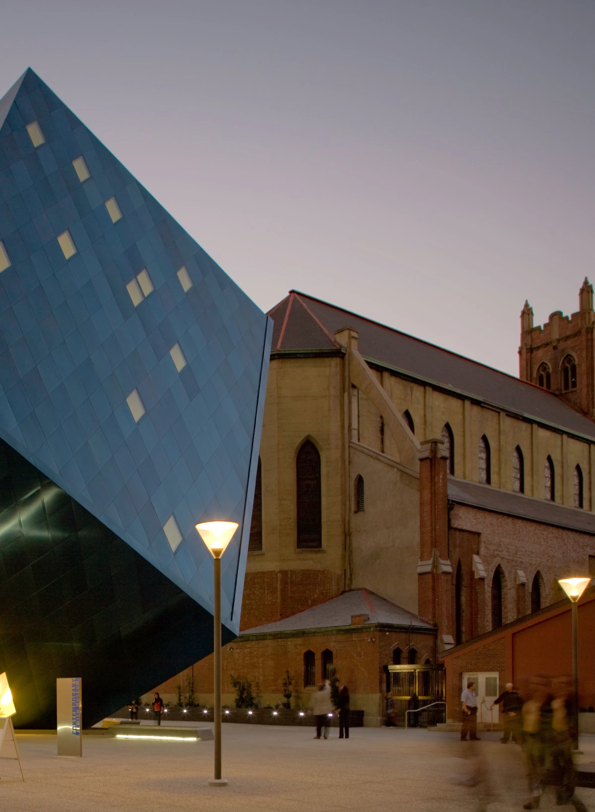 Louis Poulsen post lights illuminate the plaza beside the angular blue Jewish Museum and a historic church. © 2026 Louis Poulsen.