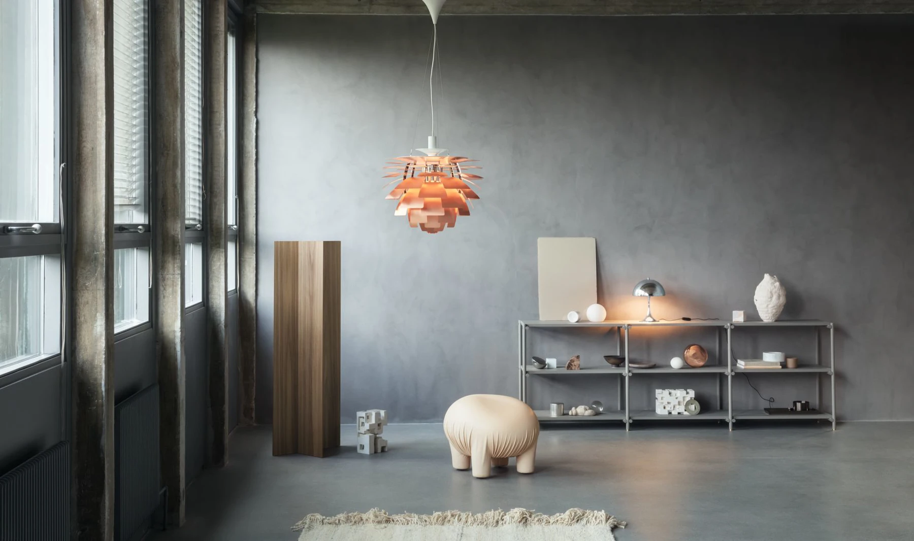 A copper PH Artichoke pendant glows above a sculptural chair in a minimalist room with grey walls. © 2026 Louis Poulsen.