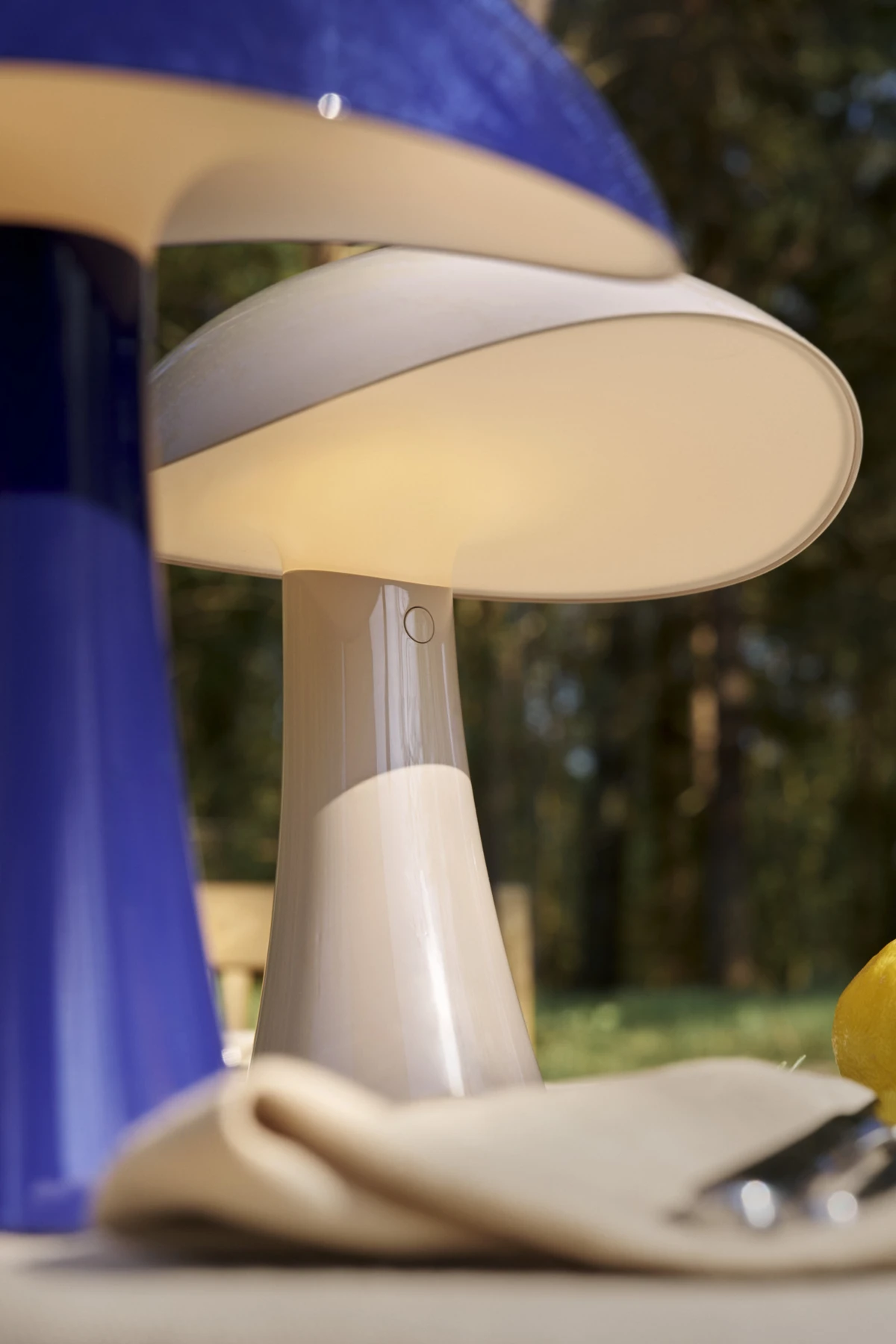 En vit Panthella 250 Portable Lamp lyser på ett utomhusbord, med en blå lampa i förgrunden. © 2026 Louis Poulsen.